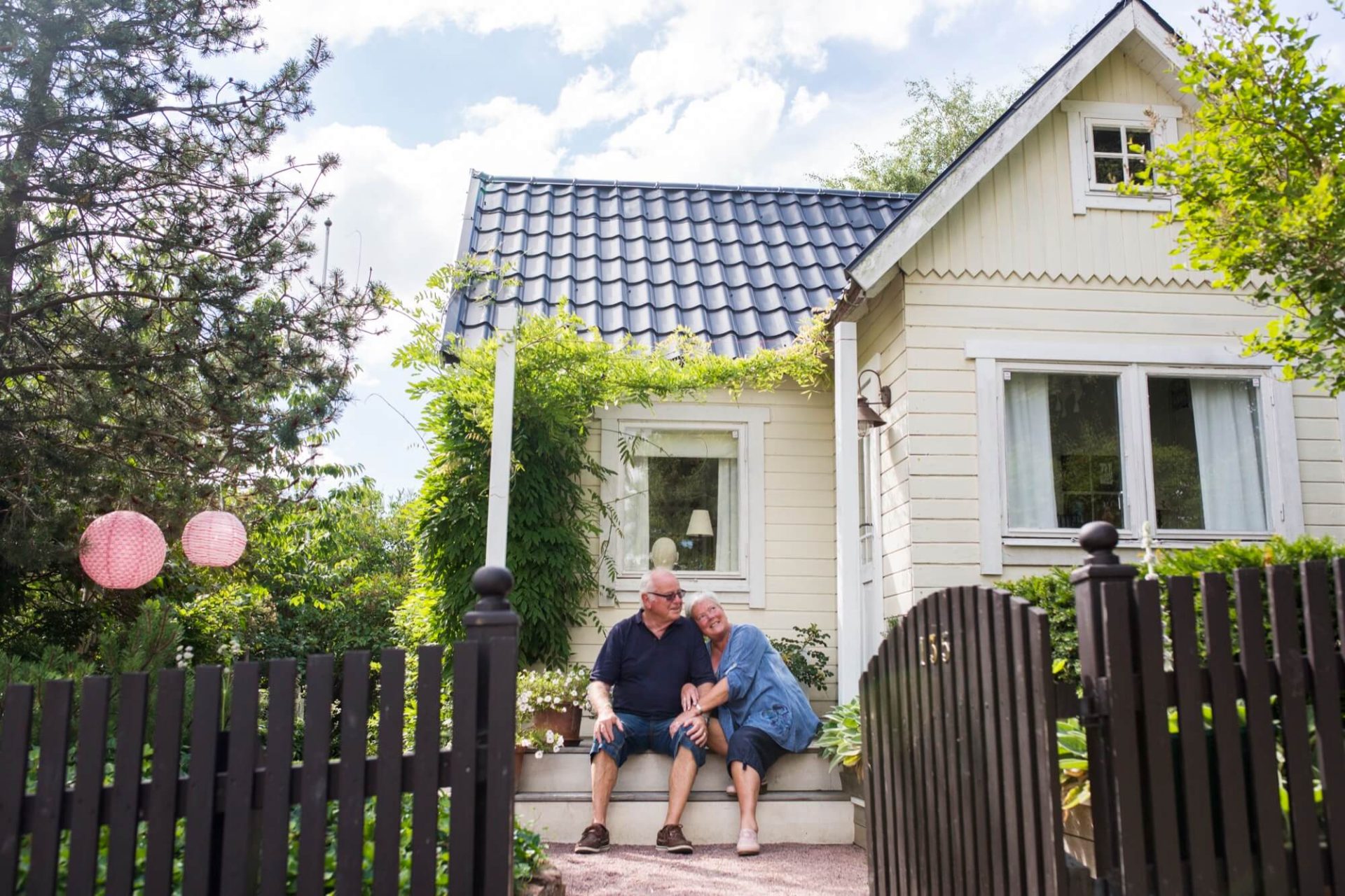 Omringat av frodig grönska, reser sig ett trähus målat i en behaglig gulton med ett mörkt tak, där vi ser ett äldre par njuta på verandan